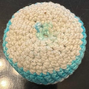 May include: A round, crocheted pincushion featuring a textured surface made of off-white yarn with a vibrant turquoise blue border. The center of the circular object displays a subtle, light blue gradient pattern integrated into the yarn stitches.