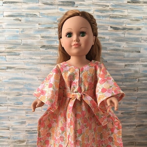 May include: A doll wearing a pink and orange floral print dress with a tie belt. The dress has long sleeves and a V-neck.