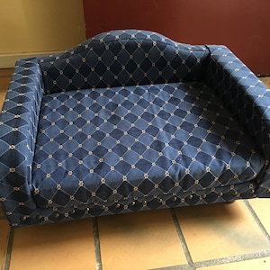 May include: A navy blue pet bed with a diamond pattern and a curved back. The bed has a cushioned interior and is trimmed with decorative studs along the base. The fabric has a repeating pattern of dark blue diamonds with small floral accents.