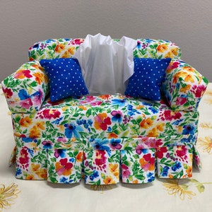 Op de afbeelding: Een tissueboxhoes met een bloemenpatroon in de vorm van een bank met blauwe kussens met witte stippen. De tissueboxhoes heeft een witte ruche aan de onderkant.