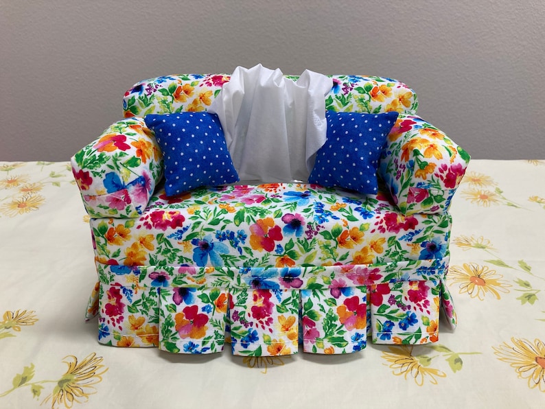 Op de afbeelding: Een tissueboxhoes met een bloemenpatroon in de vorm van een bank met blauwe kussens met witte stippen. De tissueboxhoes heeft een witte ruche aan de onderkant.