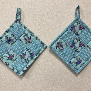 May include: Two blue quilted pot holders with a floral pattern featuring birds. The pot holders have a loop for hanging.