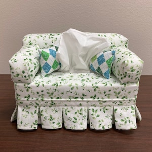 Op de afbeelding: Een witte tissueboxhoes in de vorm van een bank met een groene blad- en witte bloemenprint. De bank heeft twee kussens met een groen, blauw en wit ruitpatroon.