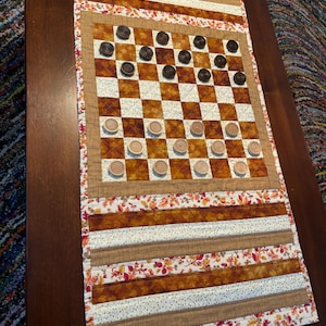 May include: A brown and white chequered game board with black and white game pieces. The board is on a brown and white quilted fabric with an autumn leaf pattern.