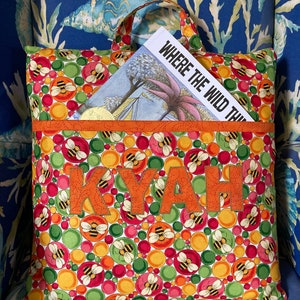 May include: A personalized book bag with a bright, colorful pattern of bees and circles in shades of red, green, yellow, and orange. The bag has a handle and a pocket on the front that says "KYAH" in orange letters. The bag is holding a book titled "Where the Wild Things Are".