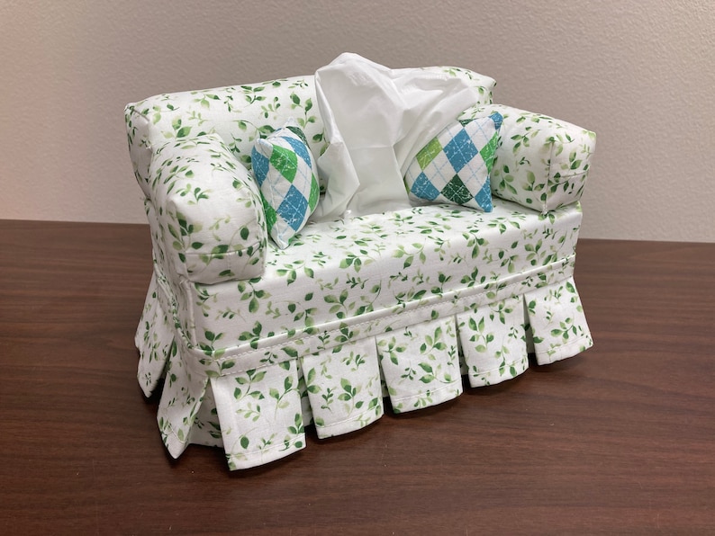 Op de afbeelding: Een witte tissueboxhoes in de vorm van een bank met groene bladerenprintstof. De bank heeft twee kussens met een groen-blauw geruit patroon.