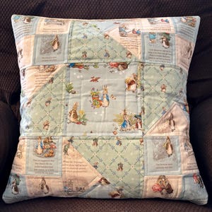 Könnte beinhalten: Quadratisches Zierkissen mit einem Patchwork-Design mit Illustrationen von Peter Hase und Text aus dem klassischen Kinderbuch. Das Kissen hat einen hellblauen Hintergrund mit einem gesteppten Muster und ist von einem braunen Rand umgeben.
