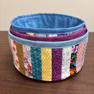 May include: A multi-coloured fabric storage basket with a blue, purple and white striped pattern. The basket is made of patchwork fabric and has a round shape.