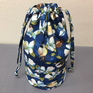 May include: A cylindrical drawstring bag with a vibrant floral and bird pattern. The fabric features white flowers, yellow and black birds, and small bird nests against a deep blue background. The bag has a drawstring closure and matching straps.