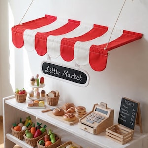Könnte beinhalten: Ein wandmontiertes Stoffvordach mit rot-weißen Streifen und einem hängenden Schild mit der Aufschrift Little Market. Unter dem Vordach befindet sich ein weißes Holzregal, gefüllt mit verschiedenen Spielzeuglebensmitteln, einer Holzkasse und einer Menütafel.