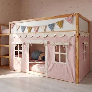 May include: A wooden loft bed frame featuring a pink fabric playhouse cover with scalloped trim, roll-up windows, and a central opening. The structure is decorated with a string of multi-coloured fabric pennant flags along the top rail.