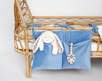 May include: A blue velvet crib organizer with two pockets, one holding a stuffed bunny and the other a wooden toy. The organizer is attached to a wicker crib.