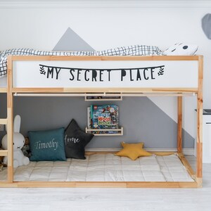 Puede incluir: Una litera de madera con un edred&oacute;n a cuadros blanco y negro en la litera superior. La cama tiene un letrero que dice "My Secret Place" sobre la litera superior. La litera inferior tiene una almohada gris y turquesa con la palabra "Timothy" en ella, una almohada gris con la palabra "Home" en ella y una almohada amarilla en forma de estrella. Hay una peque&ntilde;a cocina de juguete blanca con una estufa y un fregadero negros en el lado derecho de la imagen.