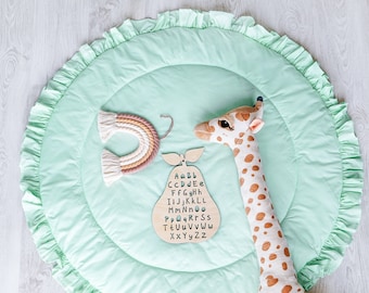 May include: A green and white play mat with a ruffled edge. The mat has a wooden pear-shaped alphabet toy and a stuffed giraffe toy on it. The pear toy has the letters of the alphabet on it.