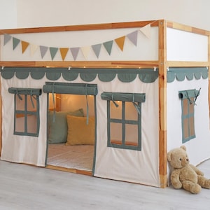 May include: A wooden playhouse-style loft bed with a white and green fabric front, complete with window features. The bed has a ladder, a small shelf, and a decorative banner. A teddy bear is placed beside the bed.