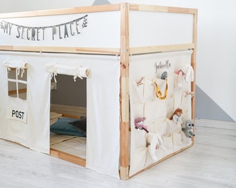 May include: A wooden bunk bed with a white canvas tent-like structure underneath. The tent has a small window with the word "POST" written on it. The bed has a white headboard with the words "MY SECRET PLACE" written on it. There are several small pockets on the side of the bed, some of which contain stuffed animals. The name "Gabriella" is written on the side of the bed.