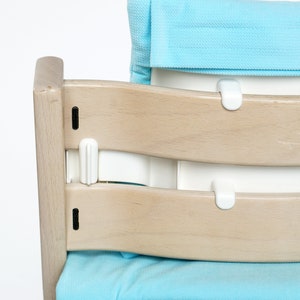 Pu&ograve; includere: Una sedia alta in legno chiaro con un sedile e uno schienale imbottiti blu. La sedia ha un cinturino in plastica bianca che attraversa lo schienale della sedia e si aggancia al sedile.