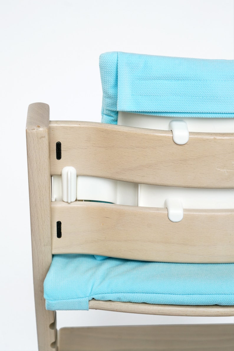 Pu&ograve; includere: Una sedia alta in legno chiaro con un sedile e uno schienale imbottiti blu. La sedia ha un cinturino in plastica bianca che attraversa lo schienale della sedia e si aggancia al sedile.