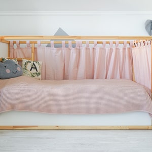May include: A wooden bed frame with pink curtains and a pink and white checkered blanket. The bed has a gray cloud pillow with a happy face and a green pillow with the letter A on it. There is a white rabbit figurine on the floor.