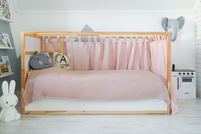 May include: A wooden bed frame with pink curtains and a pink and white checkered blanket. The bed has a gray cloud pillow with a happy face and a green pillow with the letter A on it. There is a white rabbit figurine on the floor.