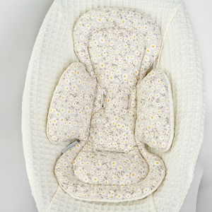 Op de afbeelding: Een witte baby schommel met een afneembare, gevoerde zittinghoes met een bloemenpatroon met kleine witte bloemen en bruine stelen op een beige achtergrond.