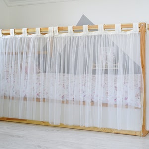 May include: A white sheer curtain canopy for a bed, with a wooden frame and white fabric ties. The canopy is attached to the top of the bed frame and hangs down to the floor.