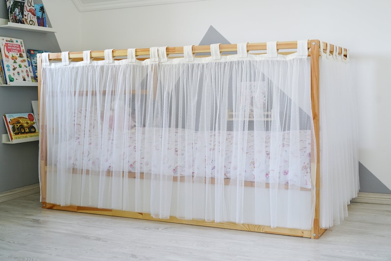 May include: A white sheer curtain canopy for a bed, with a wooden frame and white fabric ties. The canopy is attached to the top of the bed frame and hangs down to the floor.