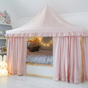 May include: A light pink fabric bed canopy with a pointed roof and ruffled trim, draped over a wooden bed frame. The canopy features long, flowing curtains that enclose the bed, which is dressed in a patterned gray and white duvet cover.