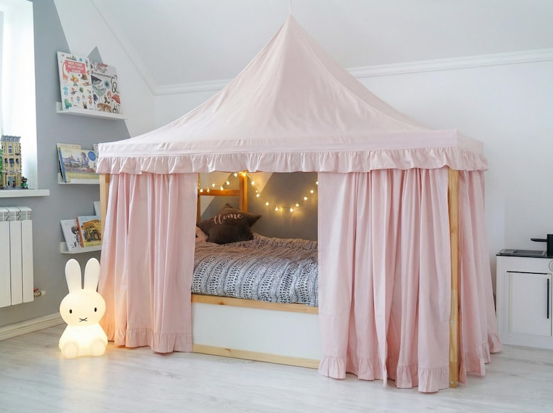 May include: A light pink fabric bed canopy with a pointed roof and ruffled trim, draped over a wooden bed frame. The canopy features long, flowing curtains that enclose the bed, which is dressed in a patterned gray and white duvet cover.