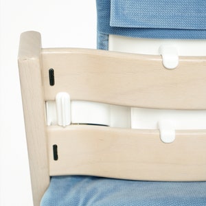 May include: A light wood high chair with a blue cushion and backrest. The chair has a curved design with white plastic safety features. The seat and backrest are upholstered in a soft, textured fabric.