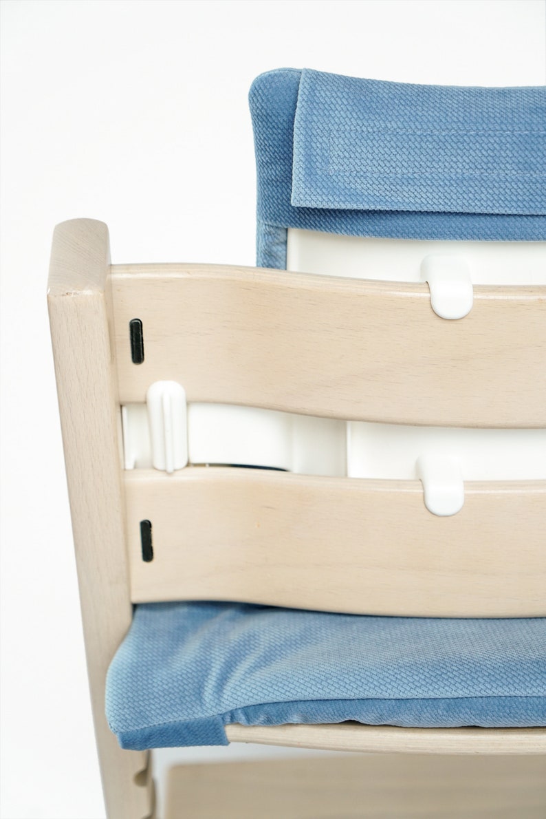 May include: A light wood high chair with a blue cushion and backrest. The chair has a curved design with white plastic safety features. The seat and backrest are upholstered in a soft, textured fabric.