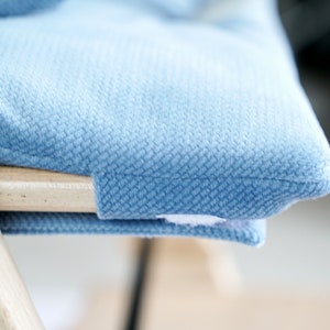 May include: Close-up of a light blue upholstered cushion on a wooden chair. The cushion has a textured surface and is attached to the chair with a small white tag. The chair's wooden frame is visible.