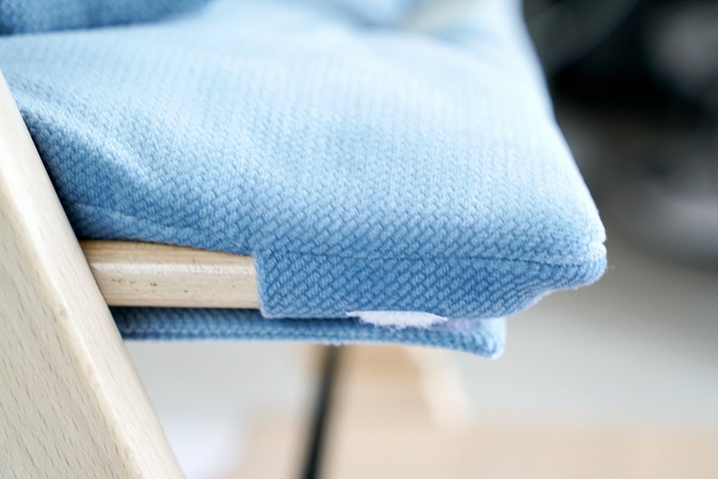 May include: Close-up of a light blue upholstered cushion on a wooden chair. The cushion has a textured surface and is attached to the chair with a small white tag. The chair's wooden frame is visible.