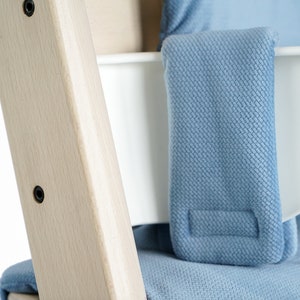May include: Close-up of a light wood high chair frame with a white seat and blue fabric padding. The frame has black screws and a light blue fabric strap. The fabric has a textured pattern.