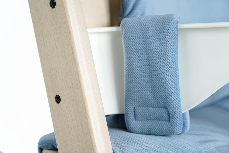 May include: Close-up of a light wood high chair frame with a white seat and blue fabric padding. The frame has black screws and a light blue fabric strap. The fabric has a textured pattern.