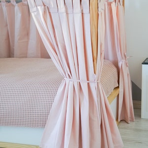May include: A pink canopy bed with a wooden frame and pink curtains. The curtains are tied back with a ribbon and the bed is made with a pink blanket.
