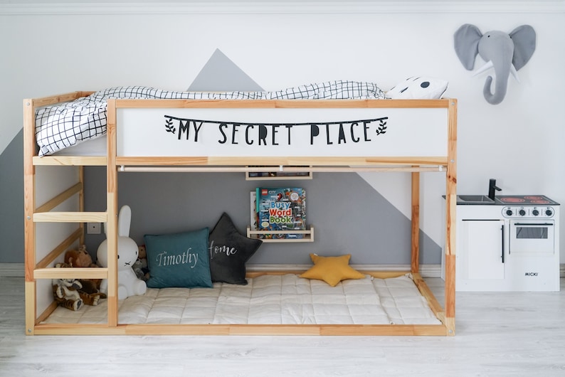 Puede incluir: Una litera de madera con un edred&oacute;n a cuadros blanco y negro en la litera superior. La cama tiene un letrero que dice "My Secret Place" sobre la litera superior. La litera inferior tiene una almohada gris y turquesa con la palabra "Timothy" en ella, una almohada gris con la palabra "Home" en ella y una almohada amarilla en forma de estrella. Hay una peque&ntilde;a cocina de juguete blanca con una estufa y un fregadero negros en el lado derecho de la imagen.