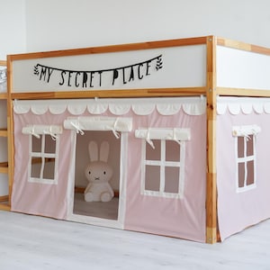May include: A wooden bunk bed with a pink fabric canopy and a white fabric lining. The canopy has scalloped edges and two small windows. The bed has a shelf on the side and a sign above the bed that says "My Secret Place".