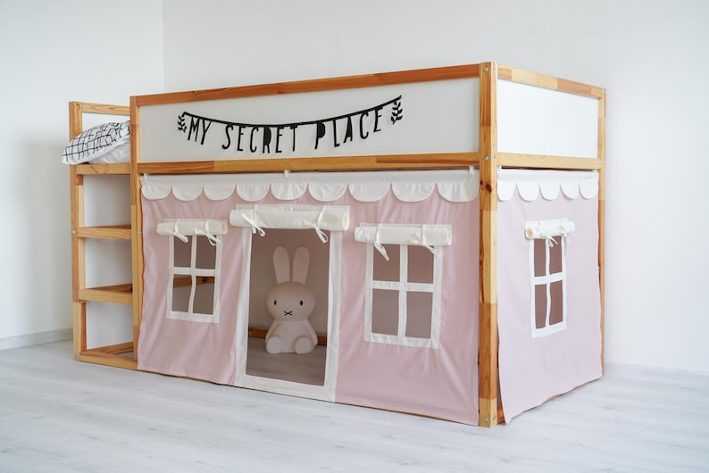 May include: A wooden bunk bed with a pink fabric canopy and a white fabric lining. The canopy has scalloped edges and two small windows. The bed has a shelf on the side and a sign above the bed that says "My Secret Place".