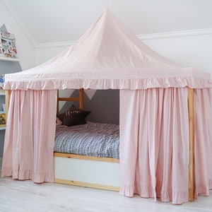 May include: A wooden bed frame featuring a light pink fabric canopy with a pointed roof and ruffled trim. The canopy drapes down to form curtains around the sides of the bed. Inside, a patterned gray and white blanket covers the mattress.