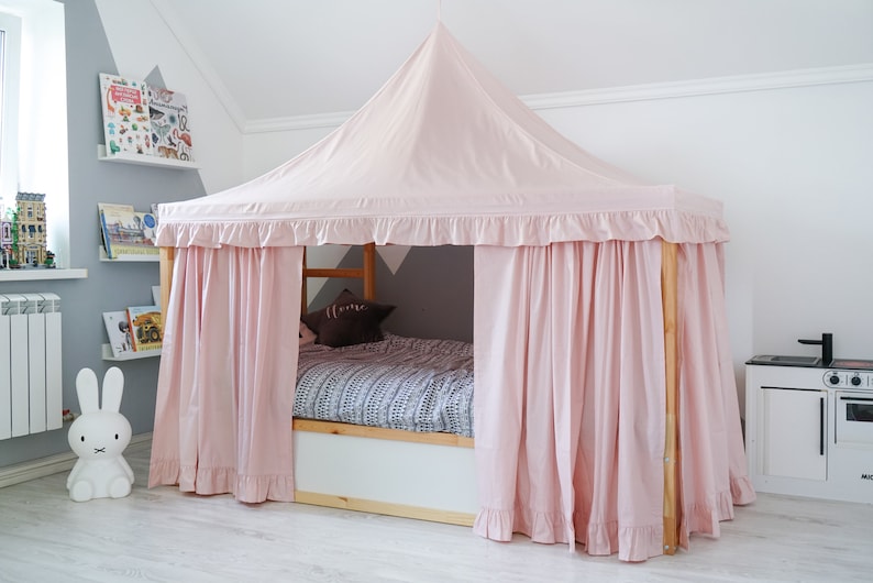 May include: A wooden bed frame featuring a light pink fabric canopy with a pointed roof and ruffled trim. The canopy drapes down to form curtains around the sides of the bed. Inside, a patterned gray and white blanket covers the mattress.