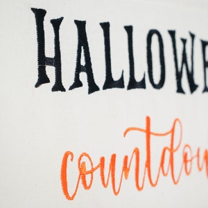 May include: Off-white Halloween countdown banner with black embroidered "Halloween" and orange "countdown" script. Features small pockets with purple ribbon trim at the bottom. Perfect for seasonal home decor.