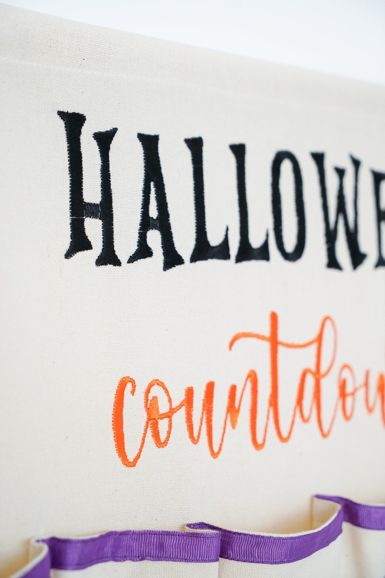 May include: Off-white Halloween countdown banner with black embroidered "Halloween" and orange "countdown" script. Features small pockets with purple ribbon trim at the bottom. Perfect for seasonal home decor.