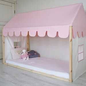 K&ouml;nnte beinhalten: Ein rosa-wei&szlig;es Himmelbettgestell mit einer gewellten Baldachin und einem wei&szlig;en Stoffpaneel mit einem Fenster. Das Bett hat eine rosa Matratze und ein wei&szlig;es Kissen mit dem Wort "CHRIST" bestickt. Auf dem Bett liegt ein Stofftier.
