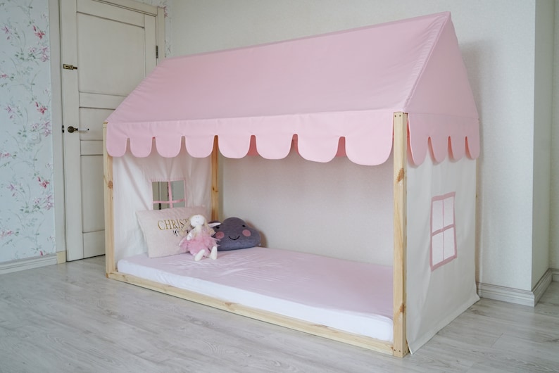 K&ouml;nnte beinhalten: Ein rosa-wei&szlig;es Himmelbettgestell mit einer gewellten Baldachin und einem wei&szlig;en Stoffpaneel mit einem Fenster. Das Bett hat eine rosa Matratze und ein wei&szlig;es Kissen mit dem Wort "CHRIST" bestickt. Auf dem Bett liegt ein Stofftier.