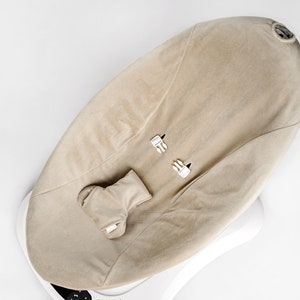 Op de afbeelding: Een beige baby wipstoel met een witte basis en een zachte, pluche stoffen bekleding. De wipstoel heeft een ingebouwde hoofdsteun en een band om de baby vast te maken.