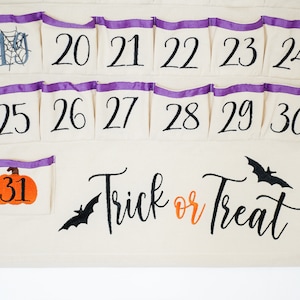 May include: A cream-colored Halloween advent calendar with numbered pockets, purple trim, and a wooden dowel. Features a pumpkin, bats, and the phrase "Trick or Treat". Includes numbers 19-31.