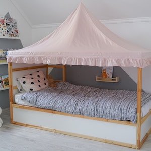 May include: A wooden bed frame with a light pink fabric canopy featuring a ruffled edge. The bed is dressed with a white pillow with black polka dots and a gray and white patterned duvet. The frame is constructed from natural light-colored wood.