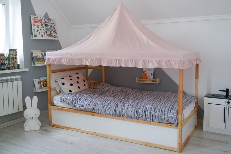 May include: A wooden bed frame with a light pink fabric canopy featuring a ruffled edge. The bed is dressed with a white pillow with black polka dots and a gray and white patterned duvet. The frame is constructed from natural light-colored wood.