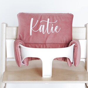 May include: A dusty rose high chair cushion set with the name "Katie" embroidered in white script. The set includes a headrest and side cushions, designed to fit a high chair. The high chair is made of light wood and has a white plastic seat.